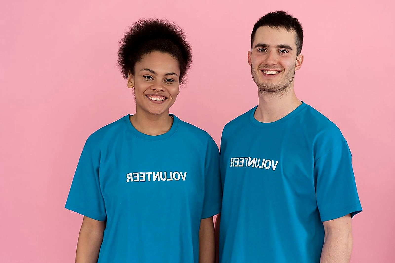 Free Two volunteers wearing t-shirts pose cheerfully against a pink studio background. Stock Photo